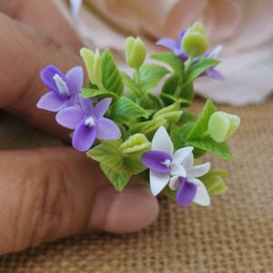 May include: A small bouquet of purple and white flowers with green leaves. The flowers are made of polymer clay and are ideal for miniature dollhouses or other craft projects.