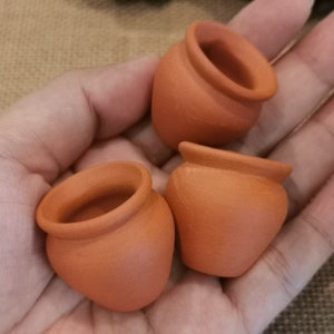 K&ouml;nnte beinhalten: Drei kleine Terrakottat&ouml;pfe, jeder mit einer leicht unterschiedlichen Form und Gr&ouml;&szlig;e, werden in einer Hand gehalten. Die T&ouml;pfe sind in einem warmen, r&ouml;tlich-braunen Ton gehalten und haben eine glatte, leicht strukturierte Oberfl&auml;che.