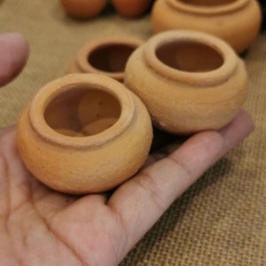 Puede incluir: Una colección de pequeñas macetas de terracota sin esmaltar. Las macetas son redondas con una abertura ancha y un borde ligeramente elevado. El color natural de la arcilla es visible. Varias macetas están enfocadas, otras borrosas en el fondo.