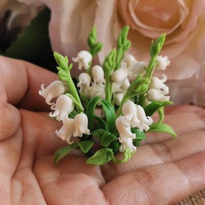 May include: A bunch of delicate white lily of the valley flowers with green stems and leaves. The flowers are arranged in a hand, creating a charming and elegant display.