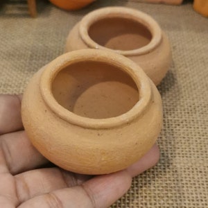 Puede incluir: Dos pequeñas macetas de terracota, una dentro de la otra, con un acabado liso y sin esmaltar. Las macetas están vacías y parecen estar hechas a mano.