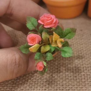 May include: Miniature arrangement of pink roses with green leaves and yellow accents in a small terracotta pot. The handcrafted floral display features three open blooms and a bud, perfect for dollhouses or craft projects.