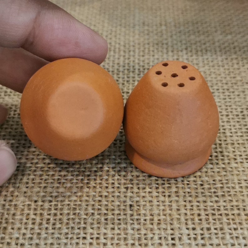 K&ouml;nnte beinhalten: Zwei Salzstreuer aus Terrakotta mit runder Basis und einer kleinen &Ouml;ffnung oben mit mehreren L&ouml;chern. Die Streuer befinden sich auf einer braunen Jutestruktur.