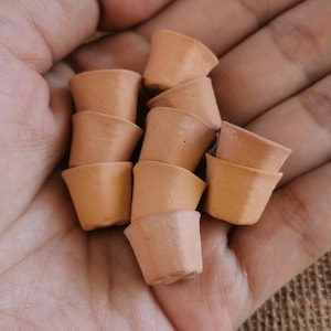 Pode incluir: Uma coleção de pequenos vasos de terracota, empilhados e dispostos em uma mão. Os vasos são de cor marrom claro e têm uma superfície lisa ligeiramente texturizada.