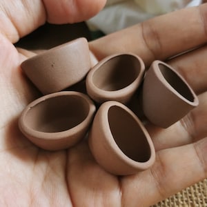 Könnte beinhalten: Fünf kleine, braune, keramische Teetassen oder Schalen sind in einer Hand angeordnet. Die Tassen sind alle in Form und Größe ähnlich, mit einem abgerundeten Boden und einem leicht ausgestellten Rand.