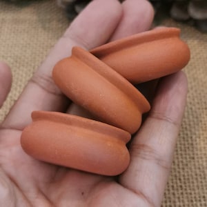 Puede incluir: Tres pequeñas macetas de terracota apiladas una encima de la otra. Las macetas son de color marrón rojizo y tienen una superficie lisa ligeramente texturizada.