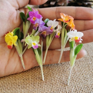 以下が含まれることがあります： 黄色、白、ピンク、紫、オレンジなど、さまざまな色の7つの造花蘭の花。花は手に置かれ、茎は下向きに伸びている。