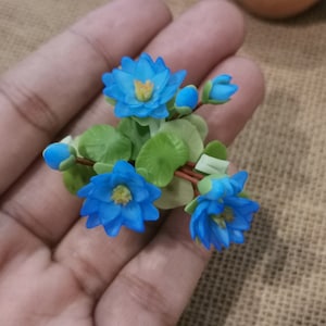 Pode incluir: Um pequeno grupo de flores azuis com folhas verdes. As flores são feitas de argila polimérica e estão dispostas em um buquê.