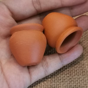 K&ouml;nnte beinhalten: Zwei kleine Terrakottat&ouml;pfe, einer mit abgerundetem Boden und der andere mit flachem Boden, sind in einer Hand zu sehen.