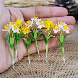 May include: Six miniature yellow and white daffodil flower stems with green leaves. The stems are white and the flowers are made of a soft material.