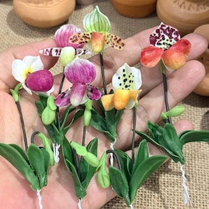 May include: A selection of handmade orchid flowers in diverse colours, such as pink, white, yellow, and red. Each flower features detailed petals and green leaves, with dark stems. The orchids are grouped together, highlighting their unique designs.
