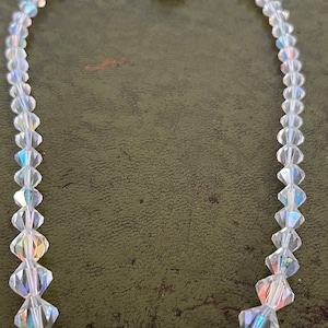 Può includere: Una collana di cristallo trasparente con sfaccettature iridescenti. La collana presenta un mix di piccole perle rotonde e cristalli più grandi, di forma geometrica. La chiusura è un piccolo pezzo argentato ornato di pietre iridescenti.