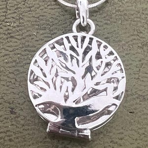 May include: Silver pendant necklace with a circular locket showcasing a tree of life design. The tree is intricately cut out, and the pendant hangs from a delicate silver chain. The locket has a hinged opening.