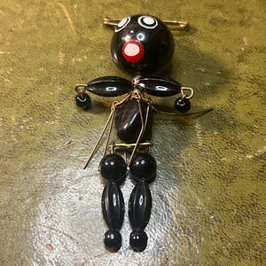 Puede incluir: Broche vintage de muñeca de cuentas negras. La muñeca tiene una cabeza redonda con ojos blancos y boca roja. Cuenta con brazos y piernas ovalados negros, y está sujeta a un imperdible dorado.