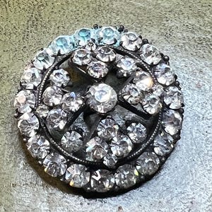 Könnte beinhalten: Eine runde, antike Brosche mit einem silberfarbenen Metallrahmen. Die Brosche ist mit zahlreichen klaren, funkelnden Strasssteinen besetzt, die kreisförmig angeordnet sind. In der Mitte befindet sich ein größerer, prominenter Stein. Die Brosche hat eine Vintage-Ästhetik.