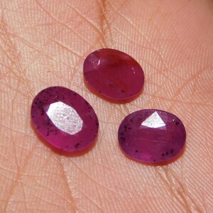 Puede incluir: Tres rubíes facetados de forma ovalada con un color rojo magenta intenso. Cada piedra preciosa presenta una superficie pulida con inclusiones oscuras visibles dispersas por el material translúcido, reflejando la luz en sus facetas planas.