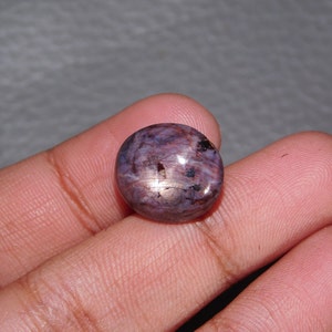 May include: A round, cabochon-cut gemstone featuring a deep purple and reddish-brown colour palette with visible internal inclusions and a distinct six-rayed star effect, known as asterism, reflecting light across its polished, smooth surface.