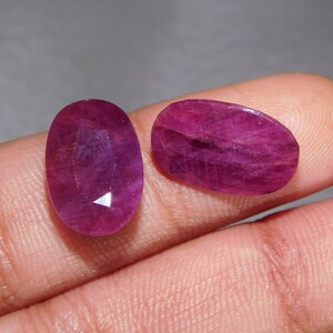 May include: Two loose, faceted gemstones with a deep magenta-red color. One stone is oval-shaped and oriented vertically, while the other is a flatter, elongated oval shape. Both stones exhibit a translucent quality with visible internal inclusions.