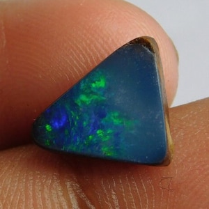 May include: A triangular-shaped dark blue opal gemstone featuring vibrant flashes of green and blue play-of-color across its polished surface. The stone has a smooth, slightly rounded edge and a deep, saturated base color with distinct iridescent inclusions.