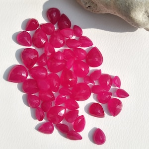 May include: A collection of numerous small, faceted, teardrop-shaped gemstones in a vibrant hot pink color. Each individual stone features a polished surface with multiple geometric facets that reflect light, creating a sparkling appearance.