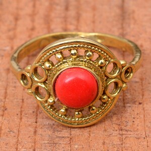 Puede incluir: Anillo dorado con una piedra roja brillante y redonda en el centro. El anillo presenta un diseño circular ornamentado alrededor de la piedra y en los laterales, creando una estética vintage o bohemia. El anillo se encuentra sobre una superficie de madera cálida.