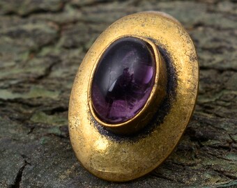 May include: A gold-toned ring with a purple gemstone set in the center. The ring is oval-shaped and has a raised edge around the gemstone.