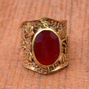 Può includere: Un anello color oro con una gemma ovale, sfaccettata, rosso scuro. L'anello ha una fascia dettagliata e strutturata con un design floreale e di foglie. L'anello è posto su uno sfondo di legno.