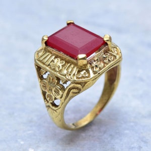Könnte beinhalten: Ein goldener Ring mit einem verzierten Design und einem großen, quadratischen roten Edelstein.
