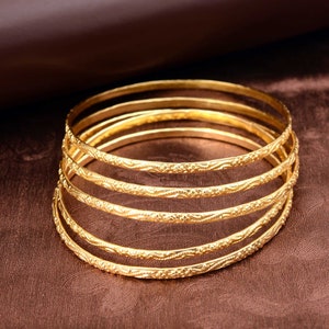 Puede incluir: Un conjunto de seis pulseras doradas con intrincados diseños grabados. Las pulseras están apiladas una encima de la otra, creando un aspecto en capas.