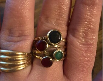 May include: Four gold rings with different colored stones. The rings are stacked on a finger. The stones are black, red, green, and red.