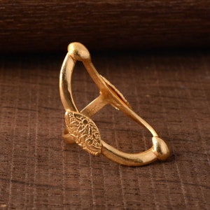 Puede incluir: Un anillo dorado con un diseño abierto. El anillo presenta un elemento decorativo ovalado con un patrón floral en relieve. La banda del anillo se curva para encontrarse en una punta redondeada, creando una forma única y llamativa.