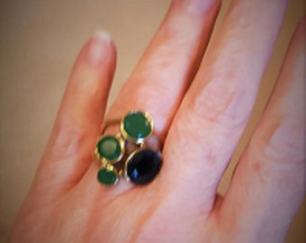 May include: A gold ring with multiple green and black gemstones. The ring is on a person's finger.