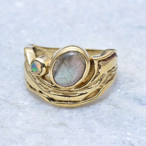 Op de afbeelding: Een gouden ring met een grote ovale labradorit-edelsteen en een kleinere ronde opaal-edelsteen. De ring heeft een getextureerd, organisch ontwerp.