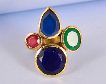 May include: Gold ring with four colorful gemstones. The ring features a large round blue stone, a smaller round red stone, a teardrop-shaped blue stone, and a round green stone.