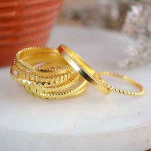 May include: A set of five gold-toned stacking rings with different textured patterns. The rings are stacked on a white surface.