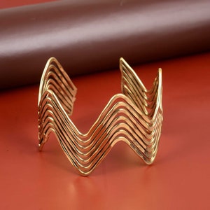 Op de afbeelding: Een goudkleurige manchetarmband met een golvend ontwerp. De open manchet heeft meerdere parallelle, golvende lijnen, waardoor een zigzagpatroon ontstaat. De armband staat tegen een rood-oranje achtergrond.