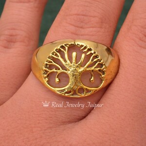 May include: A gold-colored ring featuring a detailed tree of life design. The ring has a smooth, polished band and a raised, oval-shaped face with the tree motif. The text "Real Jewelry Jaipur" is at the bottom of the image.