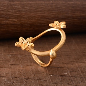 May include: A gold-colored ring with a floral design. The ring features two openwork flower accents and a small, rounded detail. The ring is set against a wooden background.
