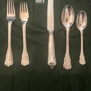 May include: A set of silver-colored flatware, including two forks, a knife, and two spoons, arranged on a dark green cloth. The flatware features ornate floral detailing on the handles. The cloth has the text "GUMPS SINCE 1861" printed in white.