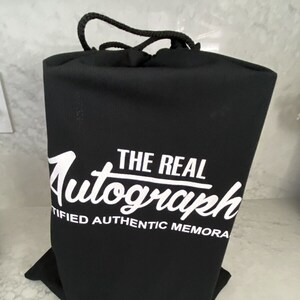 May include: A black drawstring bag with a white logo that reads "THE REAL Autograph CERTIFIED AUTHENTIC MEMORA".