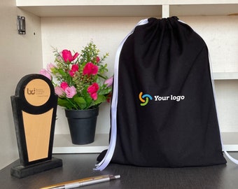 Könnte beinhalten: Schwarze Kordelzugtasche mit weißen Kordeln und einem weißen Logo mit der Aufschrift "Your logo" in einem bunten Kreisdesign. Die Tasche liegt auf einer Holzoberfläche mit einer kleinen Topfpflanze und einer Trophäe.
