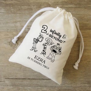 Könnte beinhalten: Ein weißer Baumwollbeutel mit Kordelzug, der eine schwarz-weiße Illustration von Woody, Jessie und Buzz Lightyear aus Toy Story zeigt. Der Beutel trägt den Text "2 infinity & beyond" und "EZRA IS TURNING TWO!" aufgedruckt.