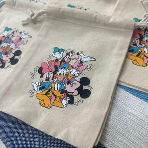 Op de afbeelding: Beige trekkoordzakken met een kleurrijke afbeelding van Disney-personages, waaronder Mickey Mouse, Minnie Mouse, Donald Duck en Goofy. De zakken hebben een klein logo aan de bovenkant en zijn gemaakt van een canvasachtig materiaal.