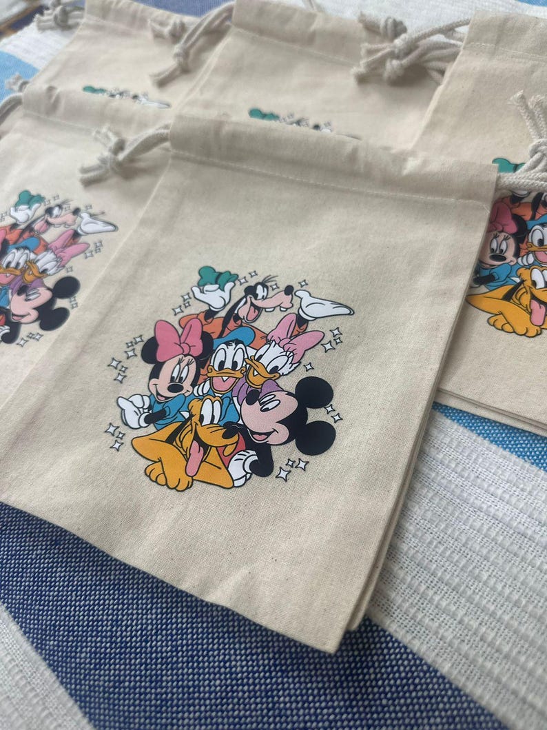 Op de afbeelding: Beige trekkoordzakken met een kleurrijke afbeelding van Disney-personages, waaronder Mickey Mouse, Minnie Mouse, Donald Duck en Goofy. De zakken hebben een klein logo aan de bovenkant en zijn gemaakt van een canvasachtig materiaal.