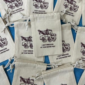 Puede incluir: Una colecci&oacute;n de peque&ntilde;as bolsas de tela blanca con cierres de cord&oacute;n marr&oacute;n. Cada bolsa presenta una imagen de dibujos animados de una gr&uacute;a roja con el texto "I got wrecked at Tow Mater Fest".