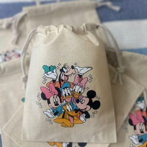 Op de afbeelding: Beige trekkoordzakken met een kleurrijke afbeelding van Disney-personages, waaronder Mickey Mouse, Minnie Mouse, Donald Duck en Goofy. De zakken zijn gemaakt van een natuurlijke stof en zijn ideaal voor feestartikelen of kleine cadeaus.