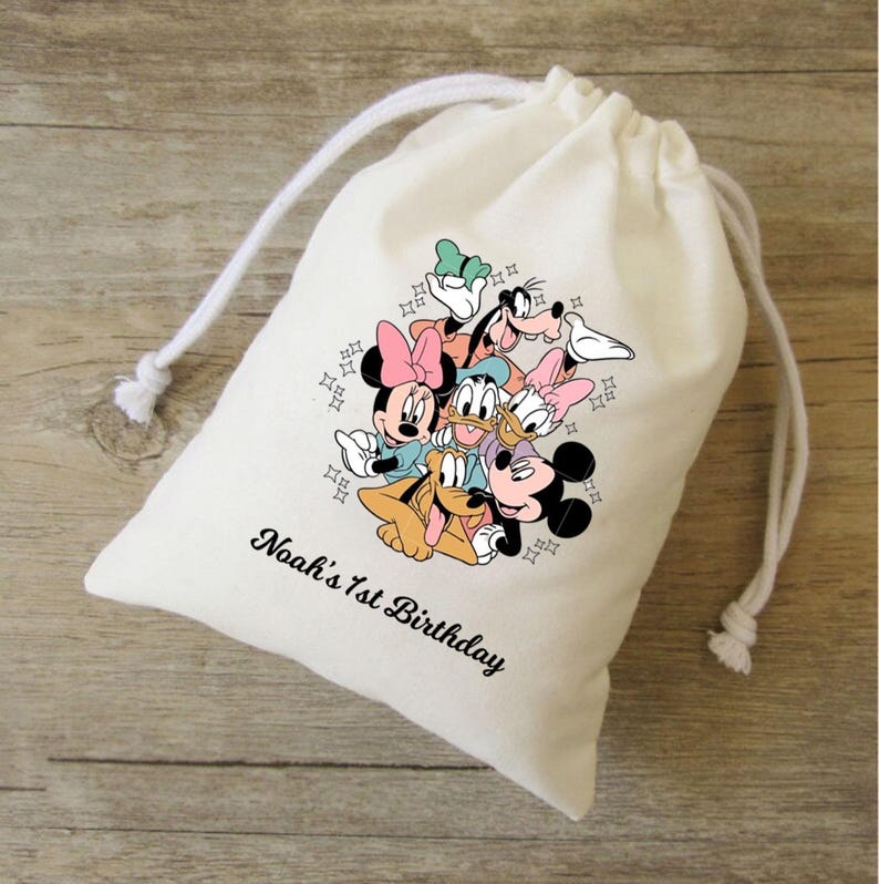 Op de afbeelding: Een witte trekkoordtas met een kleurrijke afbeelding van Disney-personages, waaronder Mickey Mouse, Minnie Mouse, Donald Duck en Goofy. De tekst "Noah's 1st Birthday" staat in zwart gedrukt. De tas is gemaakt van een zachte, lichtgekleurde stof.