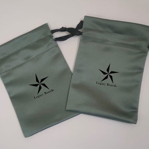 Op de afbeelding: Twee groene satijnen trekzakjes met zwarte trekkoorden en een zwart sterlogo met de tekst "Legacy Boards".