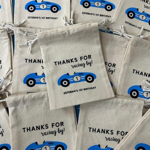 Puede incluir: Un conjunto de peque&ntilde;as bolsas de lona blancas con dise&ntilde;os de coches de carreras azules y negros. Las bolsas tienen cordones y dicen "Gracias por correr! El 1er cumplea&ntilde;os de Esteban."