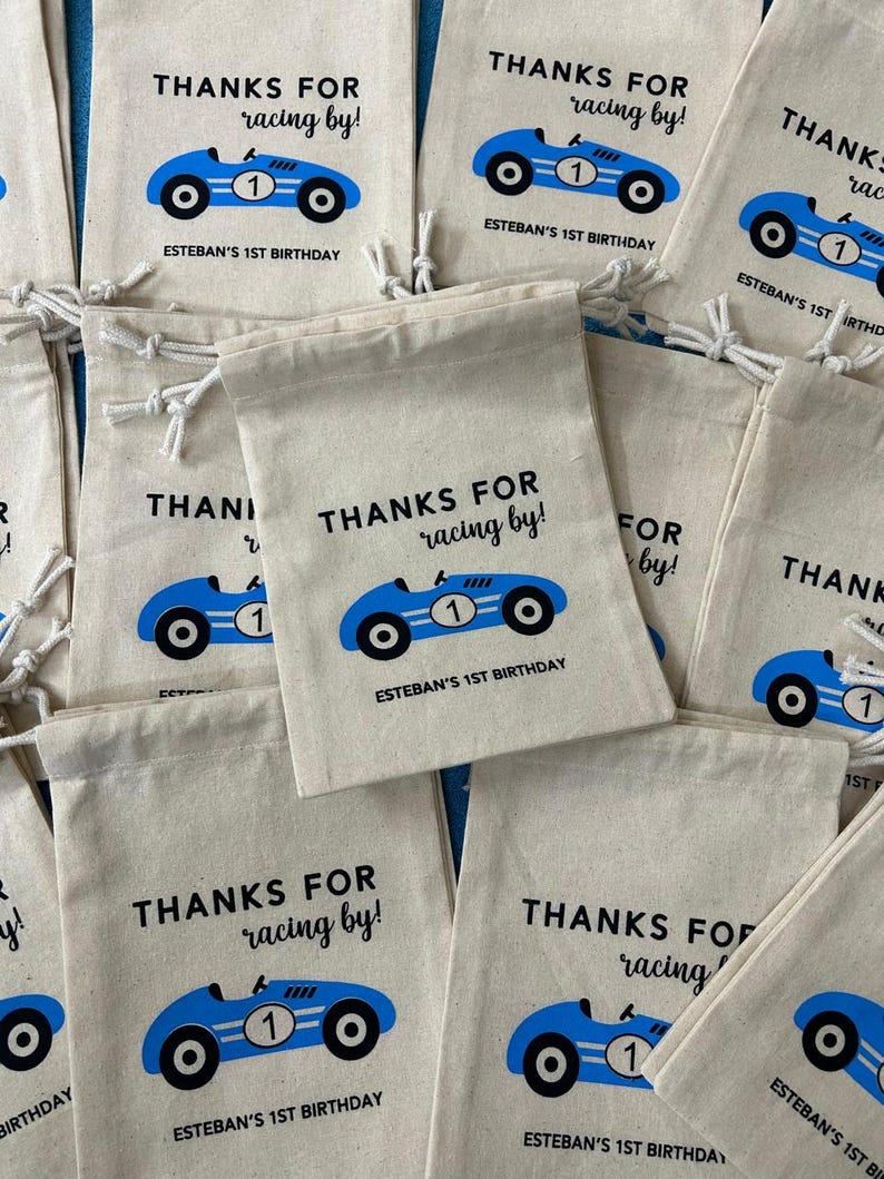 Puede incluir: Un conjunto de peque&ntilde;as bolsas de lona blancas con dise&ntilde;os de coches de carreras azules y negros. Las bolsas tienen cordones y dicen "Gracias por correr! El 1er cumplea&ntilde;os de Esteban."